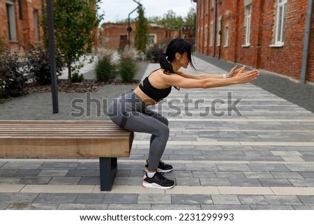 Pretty brunette woman in sportswear is doing squats while touching her buttocks to the bench outdoors Royalty-Free Stock Photo #2231299993