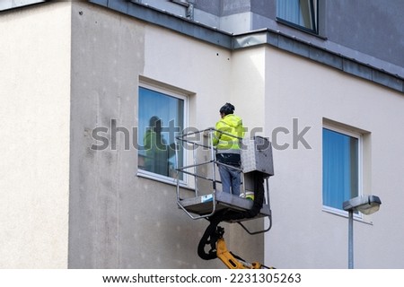 Maintenance worker in workwear using roller brush paints the facade of a residential house. Man in overalls standing outside building and repainting concrete wall close to window. Improvement concepts Royalty-Free Stock Photo #2231305263