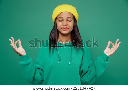 Little kid teen girl of African American ethnicity 13-14 years old wear hoody hat hold spread hand in yoga om aum gesture relax meditate calm down isolated on plain green background Childhood concept Royalty-Free Stock Photo #2231347427
