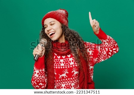 Merry fun young woman wearing red warm cozy knitted sweater hat posing sing song in microphone isolated on plain dark green background studio portrait. Happy New Year 2023 celebration holiday concept