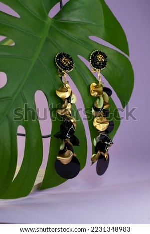 Product studio image of Long black and golden  sequins earrings clipped on green monstera leaf. Purple pink  background. Copy space for text.