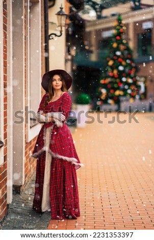 Elegant lady in vintage coat, red checkered dress and hat standing near Christmas shop with snow .Beautiful model in winter art picture. Luxurly girl in holiday mood.