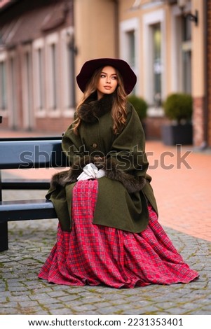 Elegant lady in vintage coat, red checkered dress and hat sitting on a bench.Beautiful model in winter art pictures. Luxurly girl in Christmas mood.