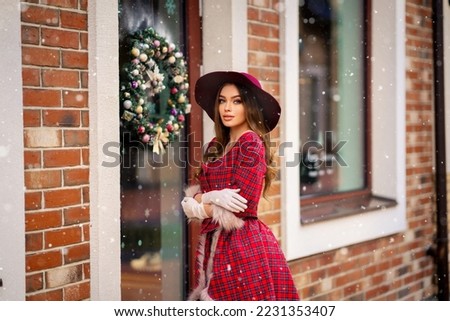 Elegant lady in vintage coat, red checkered dress and hat standing near Christmas shop with snow .Beautiful model in winter art picture. Luxurly girl in holiday mood.