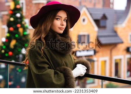 Elegant lady in vintage coat, red checkered dress and hat standing on Christmas background.Beautiful model in winter art pictures. Luxurly girl in holiday mood.