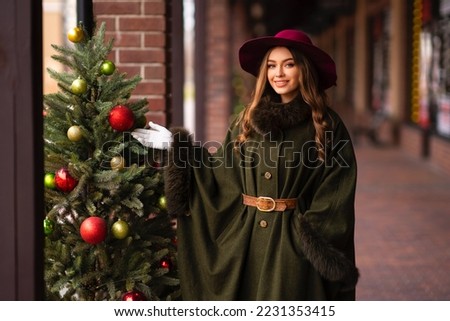 Elegant lady in vintage coat, red checkered dress and hat standing near christmas tree.Beautiful model in winter art pictures. Luxurly girl in holiday mood.