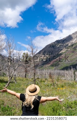 Happy woman wearing a straw hat, arms outstretched in a field, with mountain scenery. Taken in Waterton Lakes National Park Canada