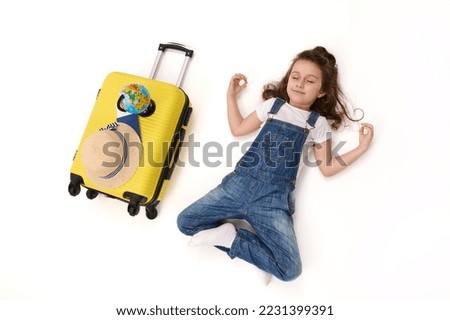 Top view of a delighted little girl in denim overalls, a traveller passenger waiting to board the flight, meditates in lotus pose, lying on white background next to her suitcase, straw hat and globe