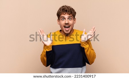 young man looking happy and excited, shocked with an unexpected surprise with both hands open next to face