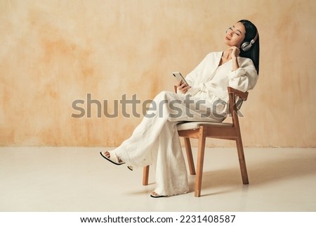 Side view of pretty asian woman sitting on chair on brown copy space background in home clothes, listening music in wireless headphones with closed eyes, using audio player app for phone Royalty-Free Stock Photo #2231408587