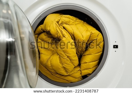 Yellow winter jacket in the drum of open washing machine in laundry room. Washing dirty down jacket in the washer Royalty-Free Stock Photo #2231408787