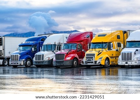 Different make big rigs semi trucks tractors with loaded semi trailers standing in the row on truck stop parking lot at early morning waiting for the route continuation time according to the log book Royalty-Free Stock Photo #2231429051