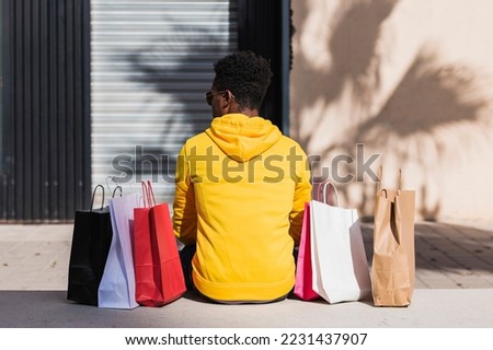 A young African man dressed in a yellow sweatshirt sitting on a street bench next to some shopping bags seen from his back.