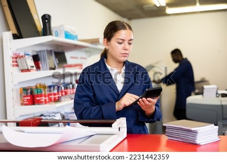Focused young female typographer in uniform calculates the cost of printed notepads and sheet in the typography
