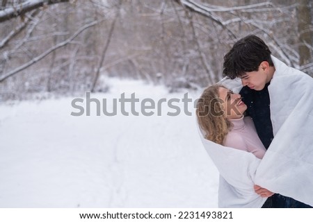 A young couple walks in the park in winter. The guy and the girl are kissing wrapped in a white blanket outdoors.