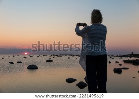 Woman taking photos in Käsmu rocky beach, Estonia. Sunset, holding a mobile phone.