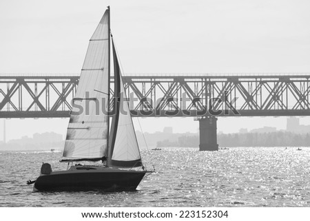 Monochrome picture of white sailing yacht catching the wind against truss bridge in autumn sunny day