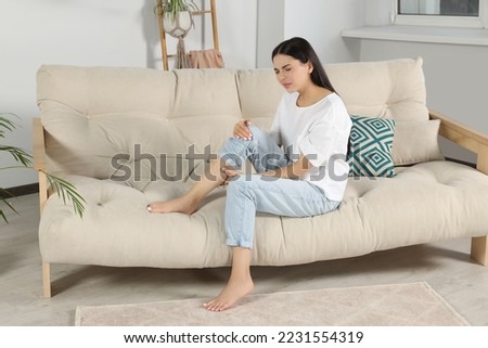 Young woman rubbing sore leg on sofa at home Royalty-Free Stock Photo #2231554319