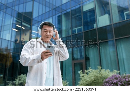 Asian man in casual clothes listening to music in headphones smiling and happy, programmer using smartphone app to listen to podcasts and online radio, man outside modern office building.