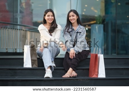 Two young women in trendy casual clothes sitting on stair in front of the shopping mall and looking at camera.