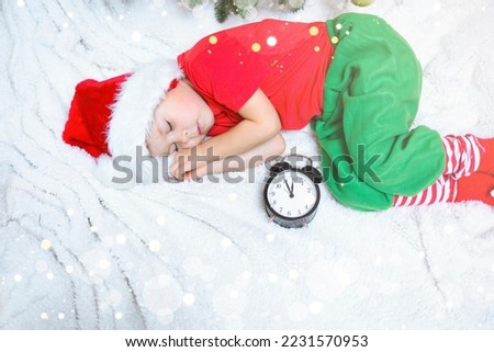 A little boy in an elf outfit in a red cap sleeps on a white blanket. The boy fell asleep under the Christmas tree with a clock, the boy is waiting for the Senta Claus. Christmas background. New Year