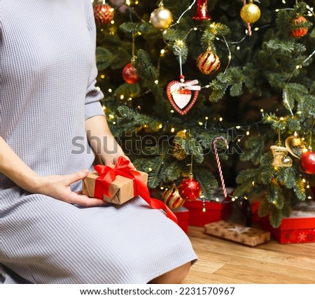 A girl in a light dress sits near the Christmas tree, holding a gift with a red ribbon. Christmas photo with gifts under the tree, celebration of the New Year and Christmas.