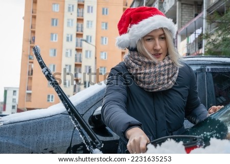 Young woman in a Santa Claus hat cleans her car from snow during the day in the courtyard of residential buildings