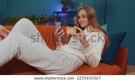 Lovely girl using credit bank card and smartphone while transferring money, purchases online shopping, order food delivery at modern home apartment indoors. Young woman in living room lying on couch