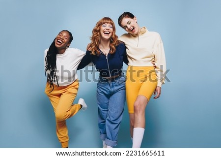 Three female friends laughing happily while embracing each other. Group of cheerful young women having fun while standing together against a blue studio background.