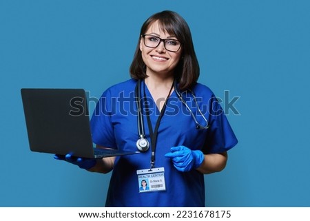 Friendly female doctor with laptop looking at camera on blue background