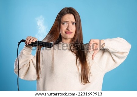 Upset young woman with flattening iron showing thumb down on light blue background. Hair damage Royalty-Free Stock Photo #2231699913