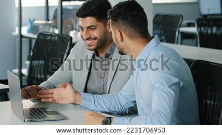 Two successful Arab male businessmen satisfied with result of negotiations, concluded contract. Smiling young colleagues Indian employees sitting in office looking at laptop screen receiving good news Royalty-Free Stock Photo #2231706953