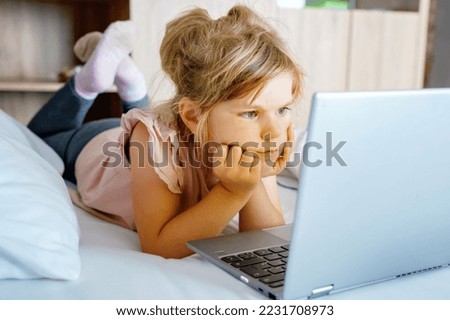 Preschool girl watching videos on laptop, notebook,in bed on clean white linens. Indoors activity with children. Freelance, distance learning or work from home with kids concept. Happy child