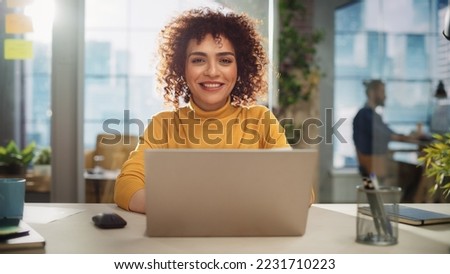 Portrait of a Beautiful Middle Eastern Manager Sitting at a Desk in Creative Office. Young Stylish Female with Curly Hair Looking at Camera with Big Smile. Colleagues Working in the Background. Royalty-Free Stock Photo #2231710223