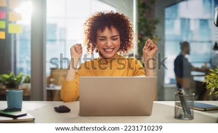 Portrait of a Beautiful Middle Eastern Manager Sitting at a Desk in Creative Office. Young Stylish Female with Curly Hair Celebrating her Achievement with Big Smile Royalty-Free Stock Photo #2231710239