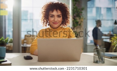 Portrait of a Beautiful Middle Eastern Manager Sitting at a Desk in Creative Office. Young Stylish Female with Curly Hair Looking at Camera with Big Smile. Colleagues Working in the Background. Royalty-Free Stock Photo #2231710301