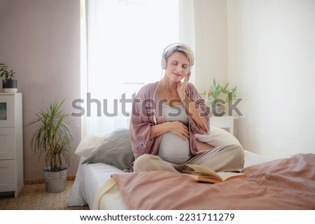 Pregnant woman sitting in her bed, listening music and reading book, enjoying time for herself.