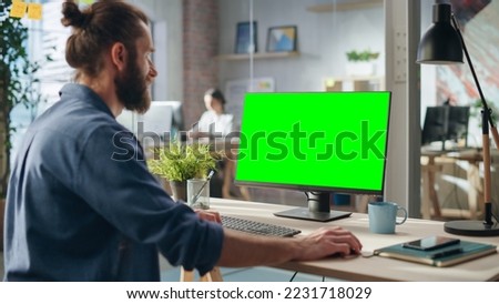 Handsome Long-Haired Bearded Manager Working at a Desk in Creative Office, Using Desktop Computer with Green Screen Mock Up Display. Colleagues Working in the Background in Marketing Agency.