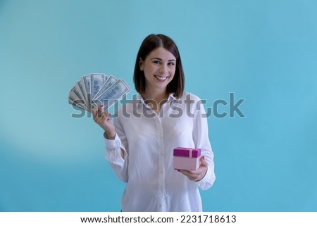 Portrait of beautiful smiling brunette woman holding pink gift box and stack of us dollars against blue background. Success theme.