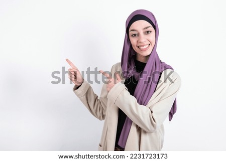 Optimistic young beautiful muslim woman wearing hijab and jacket over white background points with both hands and  looking at empty space.