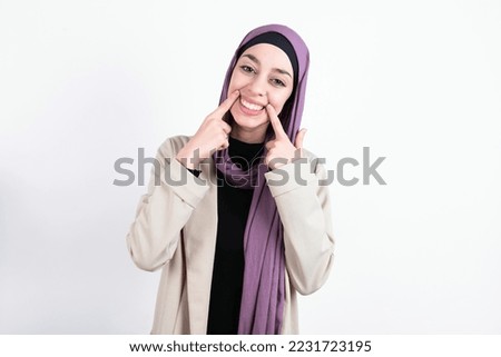 Happy young beautiful muslim woman wearing hijab and jacket over white background with toothy smile, keeps index fingers near mouth, fingers pointing and forcing cheerful smile