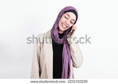 Funny young beautiful muslim woman wearing hijab and jacket over white background laughs happily, has phone conversation, being amused by friend, closes eyes.