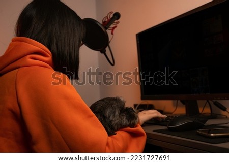 A gamer girl holding a mouse, she is using her computer and has her puppy on her lap, she is wearing an orange sweatshirt and is in her gamer room making stream.