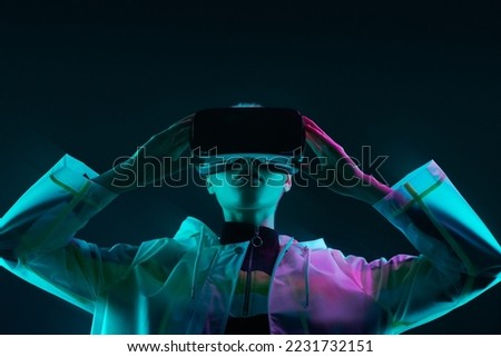 Young woman in futuristic raincoat touching VR helmet and exploring cyberspace under colorful neon light against dark background