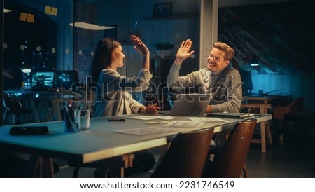 Successful Young Small Business Owners Communicating at a Meeting in Office at Night. Beautiful Female and Handsome Male Discussing Corporate Agenda and Opportunities. Managers High Five Each Other. Royalty-Free Stock Photo #2231746549
