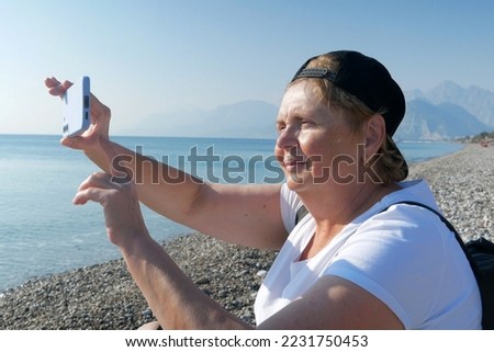 Senior adventureisageless woman with backpack taking a selfie on a mobile phone on the sea beach. Summer active tourism for pensioner. Internet for holiday concept
