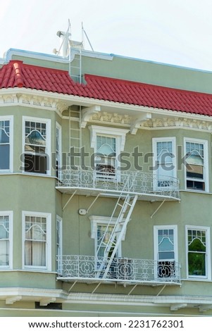Detail shot of building facade on appartment building with red roof tiles and beige paint with white fire escape ladder and flat roof. Late afternoon in shade with white and lightly blue sky.