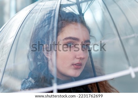 Young women with blue eyes holds transparent umbrella and looks thoughtfully through it at sky. Natural beauty