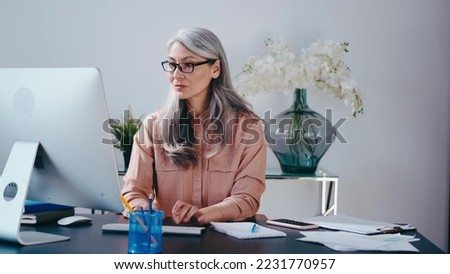 Senior stylish woman working at the computer at home. Focused cool lady freelancer writing notary in notepad.