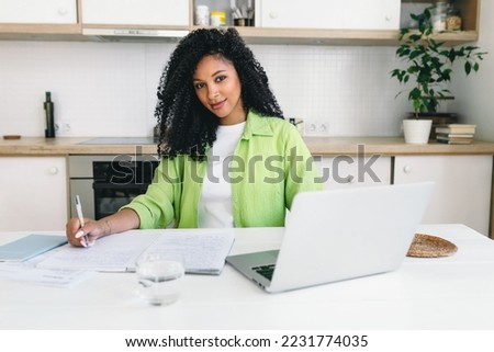 Female lawyer freelancer with dark skin and curly hair, working from home sitting at kitchen table in front of laptop, writing in her copybook, looking at camera smiling, dressed in light green shirt